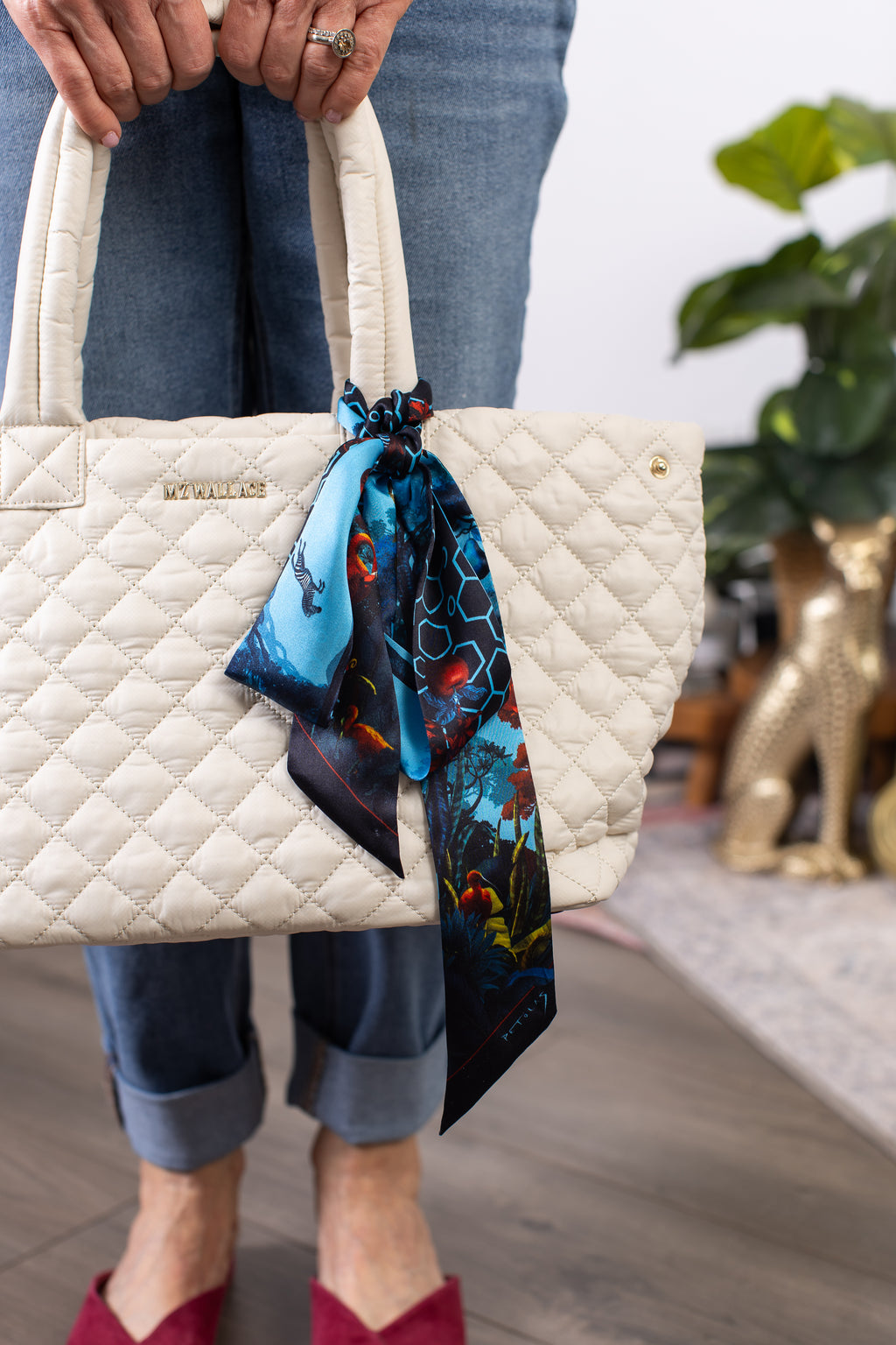 Beige quilted handbag with a colorful scarf held by a person wearing jeans and red shoes.