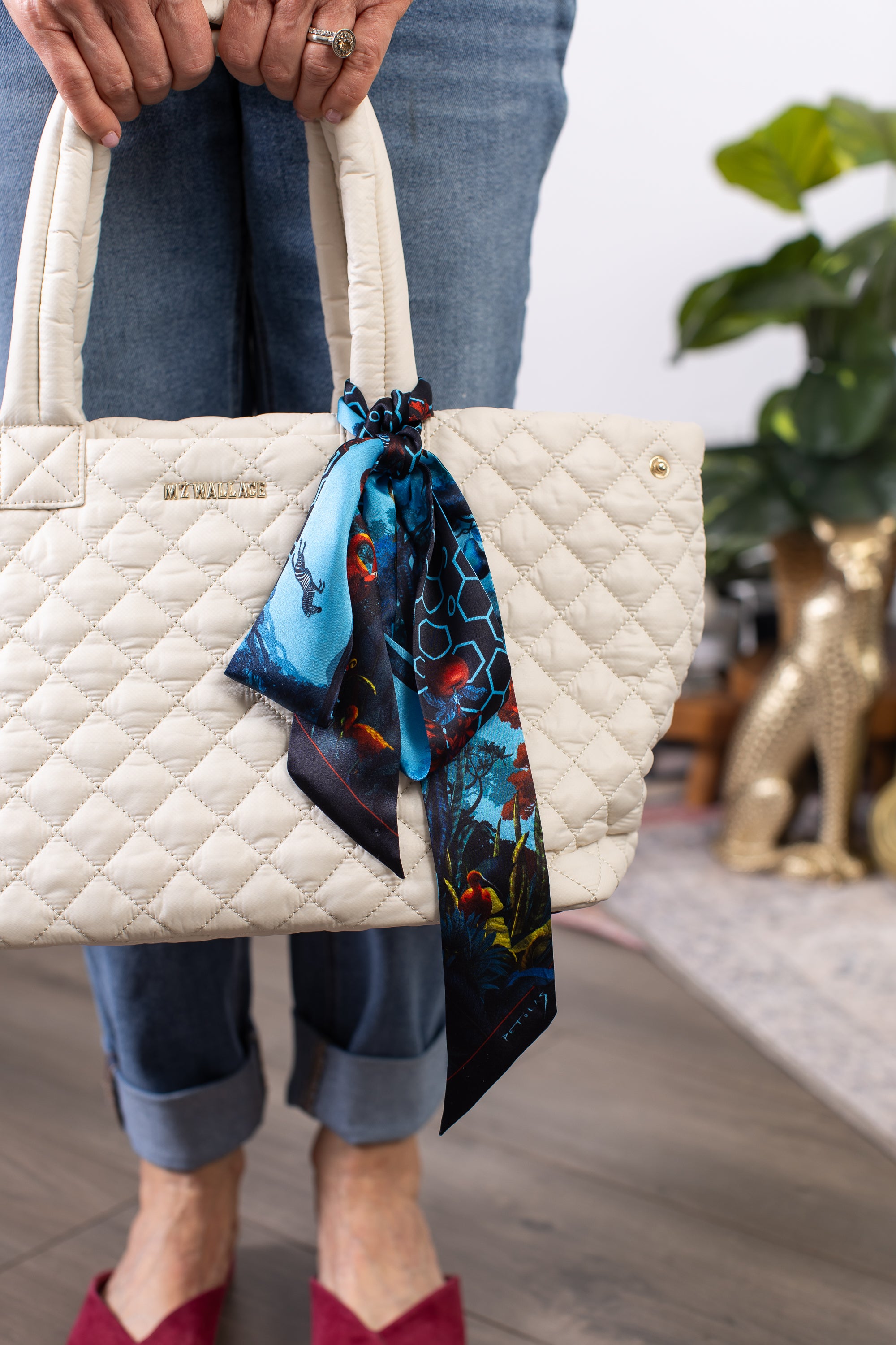 Beige quilted handbag with a colorful scarf held by a person wearing jeans and red shoes.